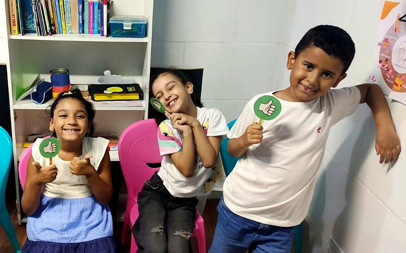 Crianças do Geração Vida sorrindo, com plaquinhas de joinha na sala do ministério infantil